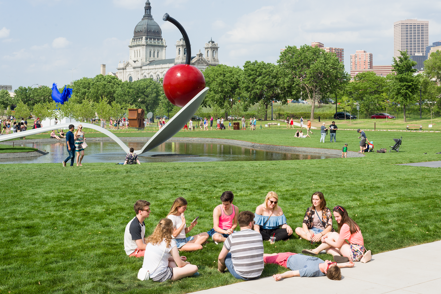 Group of people sit in a circle in front of the Spoonbridge and Cherry sculpture on a busy summer day