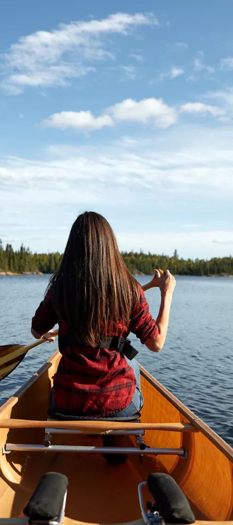 Woman in red shirt canoeing in the Boundary Waters, surrounded by dense forests and clear blue skies
